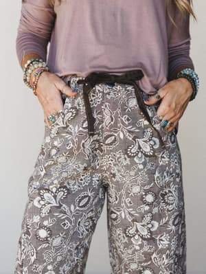 A close-cropped shot of wide-leg floral pants in a mocha-toned botanical print, styled with a dusty lavender long-sleeve top and stacked boho bracelets — the kind of outfit that looks effortless but definitely wasn't an accident.