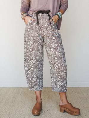 A cropped wide-leg floral pant in a warm mocha and cream botanical print, styled with a dusty mauve long-sleeve top, wood-platform clogs, and a stack of mixed bead bracelets — the kind of outfit that looks like it came together effortlessly but defin