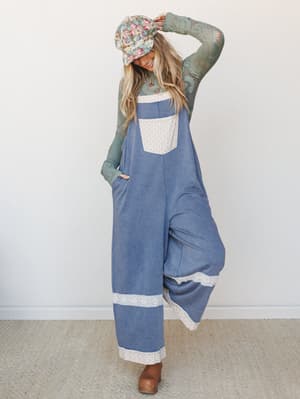 A wide-leg denim overall jumpsuit layered over a sage green lace long-sleeve top, styled with a floral bucket hat and brown clogs — the kind of effortlessly put-together look that makes people ask where you got it.