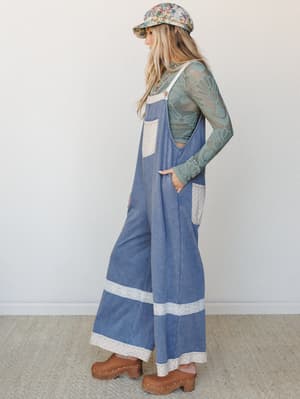 A wide-leg denim jumpsuit layered over a sage green lace top, styled with a floral newsboy cap and wood-studded clogs — the kind of outfit that feels vintage-inspired without trying too hard.