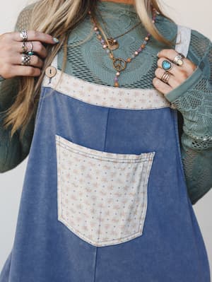 A close-up torso shot layered with a washed denim overall-style jumpsuit over a sage crochet long-sleeve top, styled with stacked boho rings and layered peace-sign necklaces — the kind of outfit that looks effortless but was absolutely thought about.