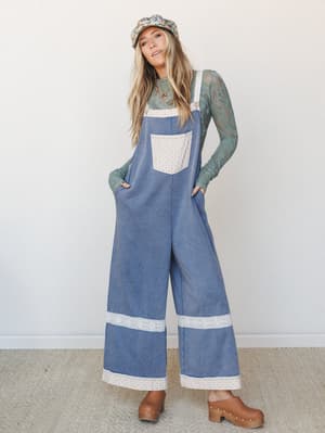 Wide-leg denim overalls with lace trim detailing styled over a sage green lace long-sleeve top, paired with a floral bucket hat and wooden clogs — the kind of outfit that feels like it was assembled on a good-mood morning.
