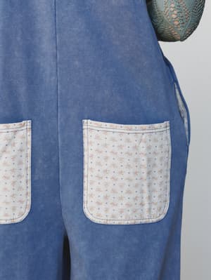 A close-up detail shot of a washed denim jumpsuit with contrast floral and polka dot patch pockets — the kind of lived-in, storybook detail that makes a piece feel personal before you even put it on.