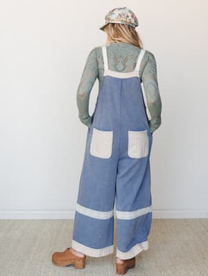 Shot from the back, this wide-leg denim overall jumpsuit is styled with a sage lace long-sleeve underneath, a floral newsboy cap, and wooden clogs — the kind of outfit that feels like it wandered in from a farmers market in the best possible way.