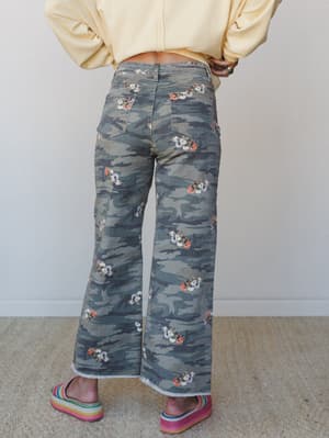 A back-view shot of wide-leg camo pants with scattered floral clusters — the kind of print that can't decide if it's tough or sweet, and honestly doesn't have to choose — paired with a butter yellow sweatshirt and rainbow flatform sandals.