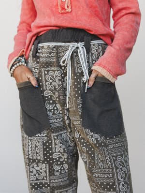 A close-up mid-shot of relaxed patchwork paisley pants in charcoal and olive tones, styled with a washed coral sweatshirt and layered bracelets — the kind of outfit that looks effortless but is clearly considered.