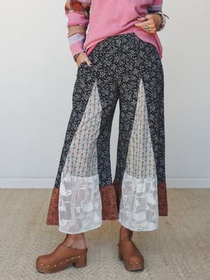 A patchwork wide-leg pant in black ditsy floral gets a serious personality boost from mixed-print insets and lace hem panels — styled with a pink thermal and wooden clogs for that effortlessly layered, lived-in boho feel.