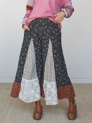 A patchwork wide-leg pant in black ditsy floral steals the show — pieced with a botanical print inset, a rust floral hem band, and lace godet panels, styled with a pink waffle knit top, stacked bracelets, and wooden clogs for a look that feels like a