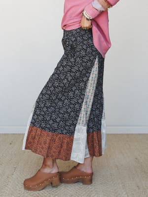 A side-profile shot of a mixed-print maxi skirt styled with a rosy pink sweatshirt and wooden clogs — the patchwork of ditsy floral, block print, and lace panels gives it that collected, well-traveled feel.
