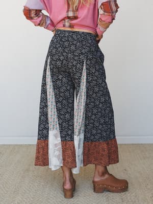 Shot from behind, a model shows off wide-leg patchwork pants in black ditsy floral with contrast panels — a boho damask inset, lace panel, and rust floral hem band — paired with a pink mixed-media top and chunky wood clogs.