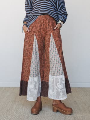 Wide-leg rust floral pants with mixed-print gusset panels, a crochet lace hem, and a dark border — styled with a striped sweater and wooden clogs for a free-spirited, pattern-mixing moment.