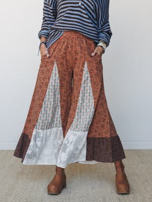 A wide-leg pant in rust floral print styled with a blue striped sweater and tan clogs — the patchwork panels, lace hem inserts, and flared silhouette give it that perfectly collected, free-spirited energy.