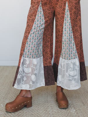 A close-up detail shot of wide-leg patchwork pants in rust floral with contrast inset panels — a geometric print meets lace hem trim — worn with wood-studded clogs on a neutral floor.