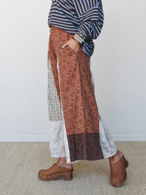 A side-profile shot of a patchwork wide-leg pant in rust with mixed floral and tile prints, lace hem panels, and a smocked waist — styled with a striped sweater, wood-soled clogs, and stacked beaded bracelets for an effortlessly layered boho look.
