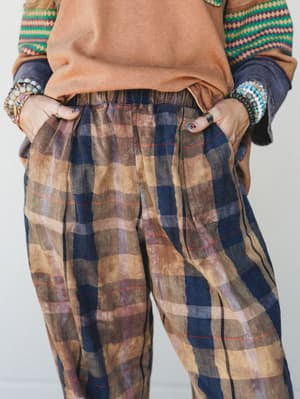 A close-up detail shot of relaxed plaid pants in earthy brown and navy, styled with a camel sweatshirt, a striped patterned layer, and a stack of mixed beaded bracelets — the kind of effortless, collected outfit that looks like it just happened.