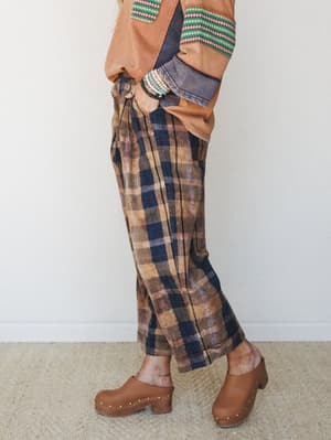 A side-profile shot of wide-leg plaid pants in washed brown and navy, styled with a rust orange top with geometric trim and worn-in clogs — the kind of outfit that looks like it came together effortlessly but absolutely didn't.