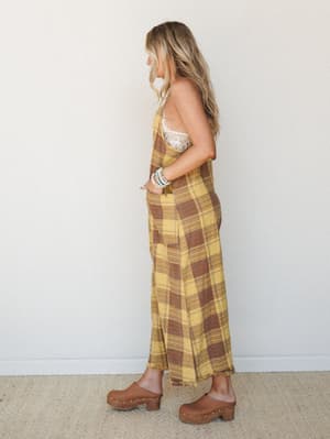 A plaid wide-leg jumpsuit in golden yellow and warm brown is styled with a lace bralette, wood-platform clogs, and stacked beaded bracelets — a vintage-leaning, free-spirited look shot in profile against a clean studio wall.
