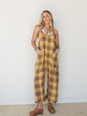 A wide-leg plaid jumpsuit in golden yellow and warm brown styled with a lace bralette, stacked bracelets, and wood clogs — effortlessly lived-in with a cool retro ease.