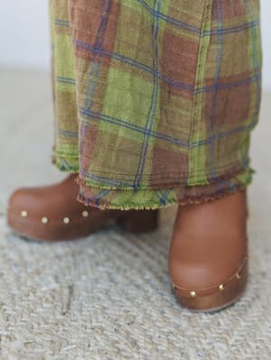 A close-up detail shot of wide-leg plaid pants with frayed hem edges, paired with tan leather platform clogs with gold stud hardware — earthy, relaxed, and very much the kind of outfit that makes a jute rug feel intentional.