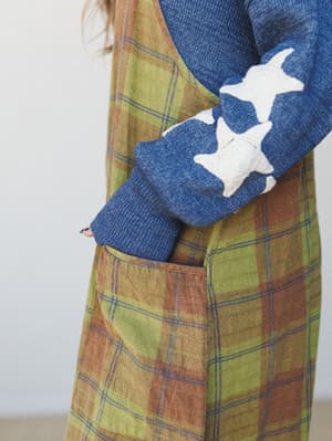 A close-up detail shot of an olive and rust plaid jumpsuit layered over a denim blue knit sweater with cream star appliqués along the sleeve — earthy, textured, and quietly expressive.
