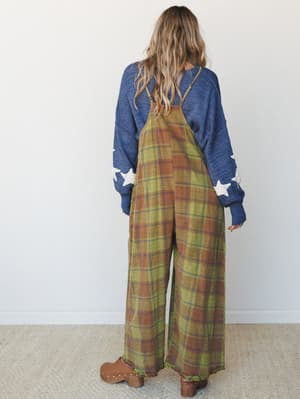 Shot from behind, she's wearing a wide-leg plaid jumpsuit in olive and rust layered over a blue star-patch sweater — the kind of effortlessly thrown-together outfit that somehow looks completely intentional.