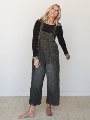 A laughing blonde model wears wide-leg denim overalls in a washed ash charcoal with a subtle leopard print overlay and distressed detailing, layered over a dark brown long-sleeve top and stacked with turquoise and beaded bracelets — the whole look ha