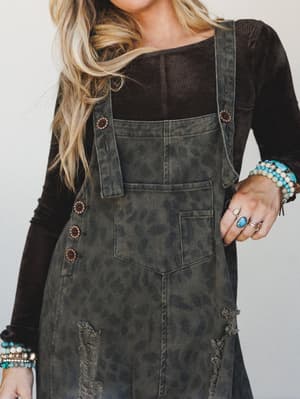 A close-up lifestyle shot of washed olive denim overalls with a faded leopard print layered over a black long-sleeve top, styled with an armful of turquoise and beaded bracelets and stacked rings — the kind of outfit that looks effortless but took ex