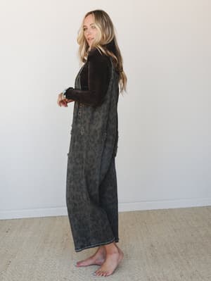 A full-length side profile shot of a model in wide-leg leopard print denim overalls layered over a dark chocolate velvet long-sleeve top, styled barefoot with a turquoise bracelet — effortlessly lived-in and a little unexpected.