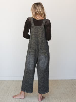 Shot from behind, a model wears wide-leg leopard print denim overalls in a washed olive-ash tone layered over a dark chocolate velvet long-sleeve top — the kind of outfit that looks effortless but is clearly very intentional.
