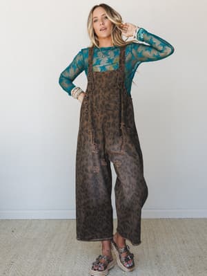 A relaxed full-body shot of a model in wide-leg leopard print overalls layered over a sheer teal lace top — the kind of outfit that looks effortless but is definitely a whole personality.