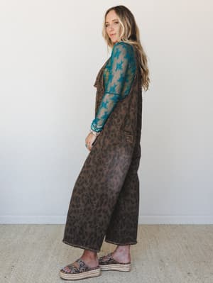 A side-profile shot of a model in wide-leg leopard print denim overalls layered over a teal lace mesh long-sleeve top — the kind of outfit that feels effortlessly collected, like she got dressed by instinct and it just worked.