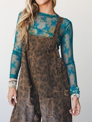 A layered boho outfit built around distressed leopard-print denim overalls worn over a sheer teal lace long-sleeve top, stacked with beaded bracelets and a delicate pendant necklace — the kind of mix-and-match that looks effortless but took real thou