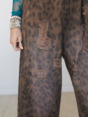 A close-up detail shot of wide-leg leopard print denim overalls in warm brown, styled with a teal lace top and an armful of layered bracelets — the distressed rips and animal print feel intentional, not accidental.