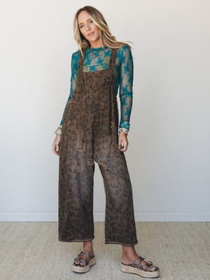 A full-body model shot of wide-leg leopard print denim overalls in washed brown, styled over a teal floral mesh long-sleeve top with platform espadrille sandals — the kind of outfit that looks effortless but clearly took some thought.