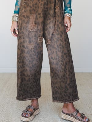 A close-up mid-shot of wide-leg leopard print denim overalls in worn brown, styled with a teal embroidered long-sleeve underneath and leopard print platform sandals — the kind of outfit that has a whole personality before you even say a word.