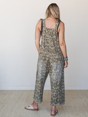 Shot from the back, a model wears wide-leg leopard-print denim overalls in a washed mushroom tone, styled with stacked turquoise bracelets and flat sandals — effortless and a little wild in the best way.