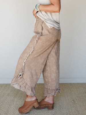 A side-profile shot of wide-leg pants in a washed almond tone with a delicate floral shadow print, styled with a cream tee, wood-studded clogs, and turquoise bead bracelets — the kind of outfit that feels effortless but clearly thought through.