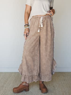 Wide-leg ruffle pants in a washed almond tone with an allover ditsy floral print, styled with a cream tee, stacked bracelets, and wood clogs — the kind of outfit that feels effortless but definitely wasn't an accident.