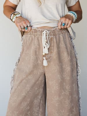 A close-up torso shot of wide-leg floral pants in a warm almond wash, styled with a cropped cream tee and stacked boho jewelry — the tassel drawstring and ruffle side seams do all the talking.