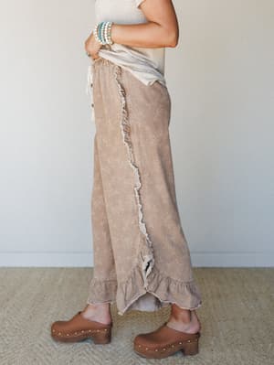 A side-profile shot of wide-leg floral pants in a warm almond tone, styled with a tucked cream tee and wood-soled clogs — the kind of outfit that feels effortless but clearly thought through.