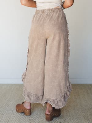 A back-view shot of wide-leg ruffle pants in a washed almond tone with a subtle floral print, paired with brown wood-sole clogs — relaxed, feminine, and the kind of thing you wear when you want to feel put-together without trying too hard.