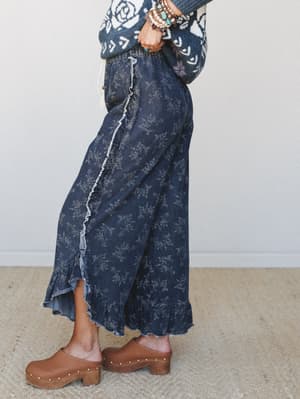 A side-profile shot of a dark denim wide-leg pant with an allover ditsy floral print, ruffle and raw-edge trim running down the side seam and hem, styled with a floral knit sweater, stacked boho bracelets, and studded wood clogs — the kind of outfit