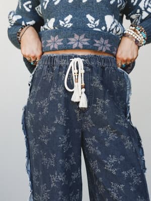 A close-up mid-shot of wide-leg floral print pants in washed dark denim, styled with a cropped fair isle sweater and layered boho jewelry — the tassel drawstring and frayed side seams give it that lived-in, effortlessly expressive feel.