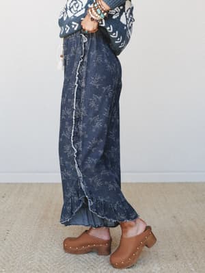 A side-profile shot of a floral-printed wide-leg pant in dark denim, styled with a cozy fair isle sweater and brown studded clogs — the kind of outfit that feels intentionally relaxed and a little bit vintage.