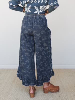 A back-view shot of wide-leg floral pants in washed dark denim with ruffle hem trim, styled with a floral jacquard sweater and chunky wood-platform clogs — the kind of outfit that feels effortless but clearly thought through.