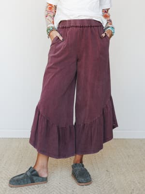 Wide-leg ruffle-hem pants in a washed mulberry hue, styled with a white tee, floral long-sleeve layer, stacked turquoise jewelry, and charcoal suede mules — relaxed and expressive with a boho-casual ease.