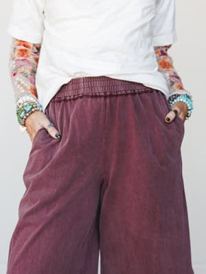 A close-up torso shot of wide-leg pants in a faded mulberry wash, styled with a white tee, floral mesh sleeves, and stacked turquoise and gemstone bracelets — relaxed and expressive in equal measure.