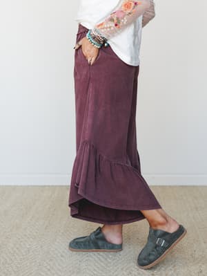 A side-profile shot of a relaxed maxi-length pant in washed mulberry, styled with a floral embroidered mesh-sleeve top, stacked bracelets, and suede buckle mules — the kind of outfit that looks effortless but clearly wasn't an accident.