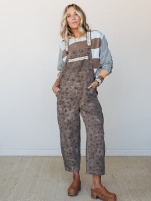 A relaxed full-body studio shot of floral-print brown overalls styled over a colorblock stripe long-sleeve top with wood clogs — the kind of outfit that feels intentional without trying too hard.