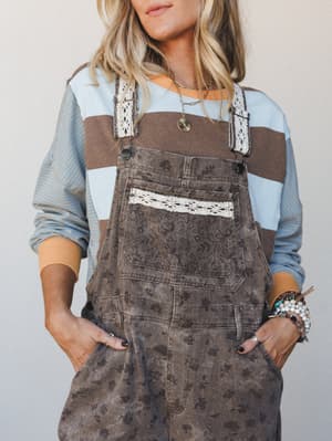 A washed brown floral-print overall layered over a color-block striped long-sleeve top, styled with stacked bracelets and layered gold necklaces — the kind of effortless, put-together look that feels intentional without trying too hard.